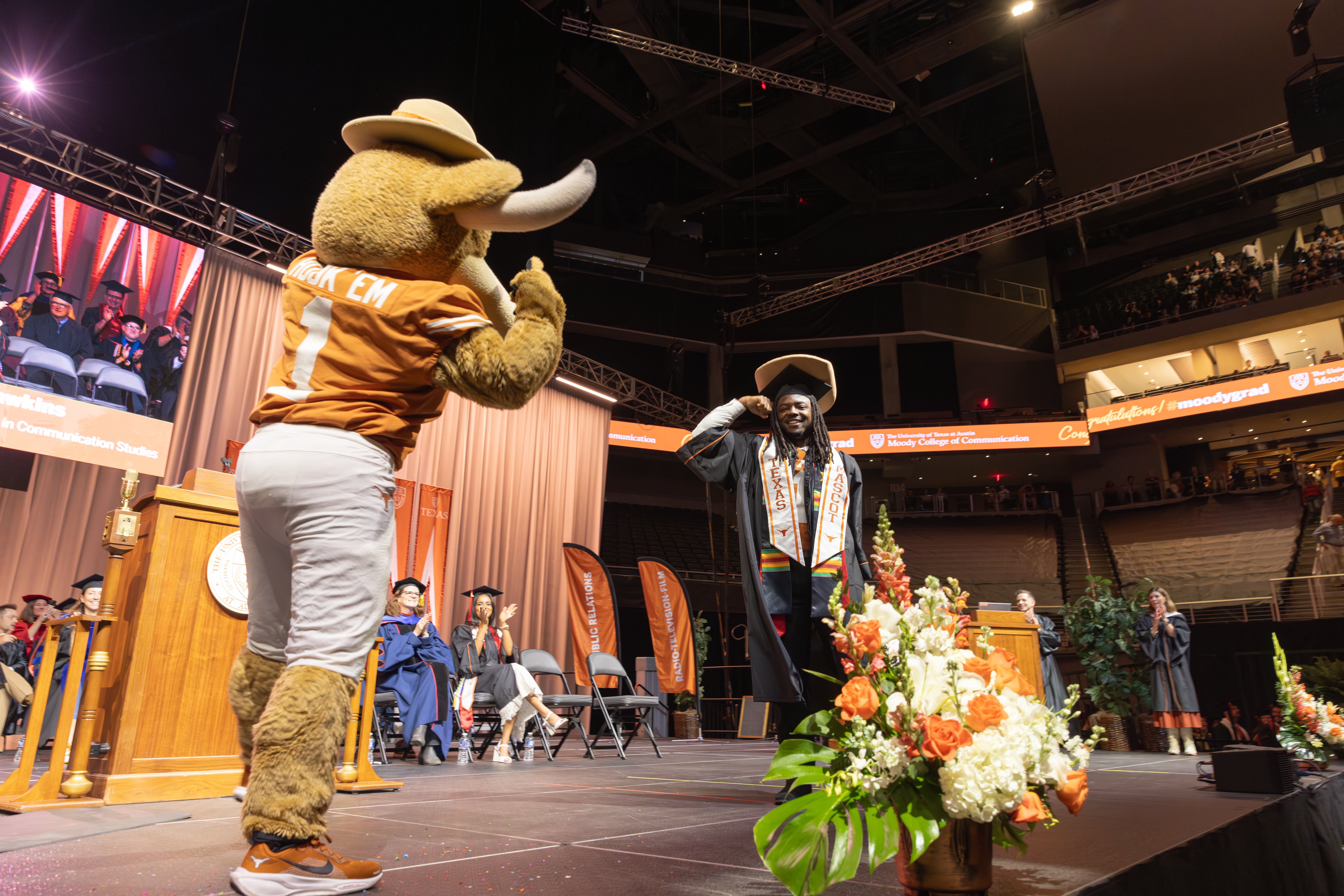 Moody College graduating student and Hook Em' walking on the graduation stage