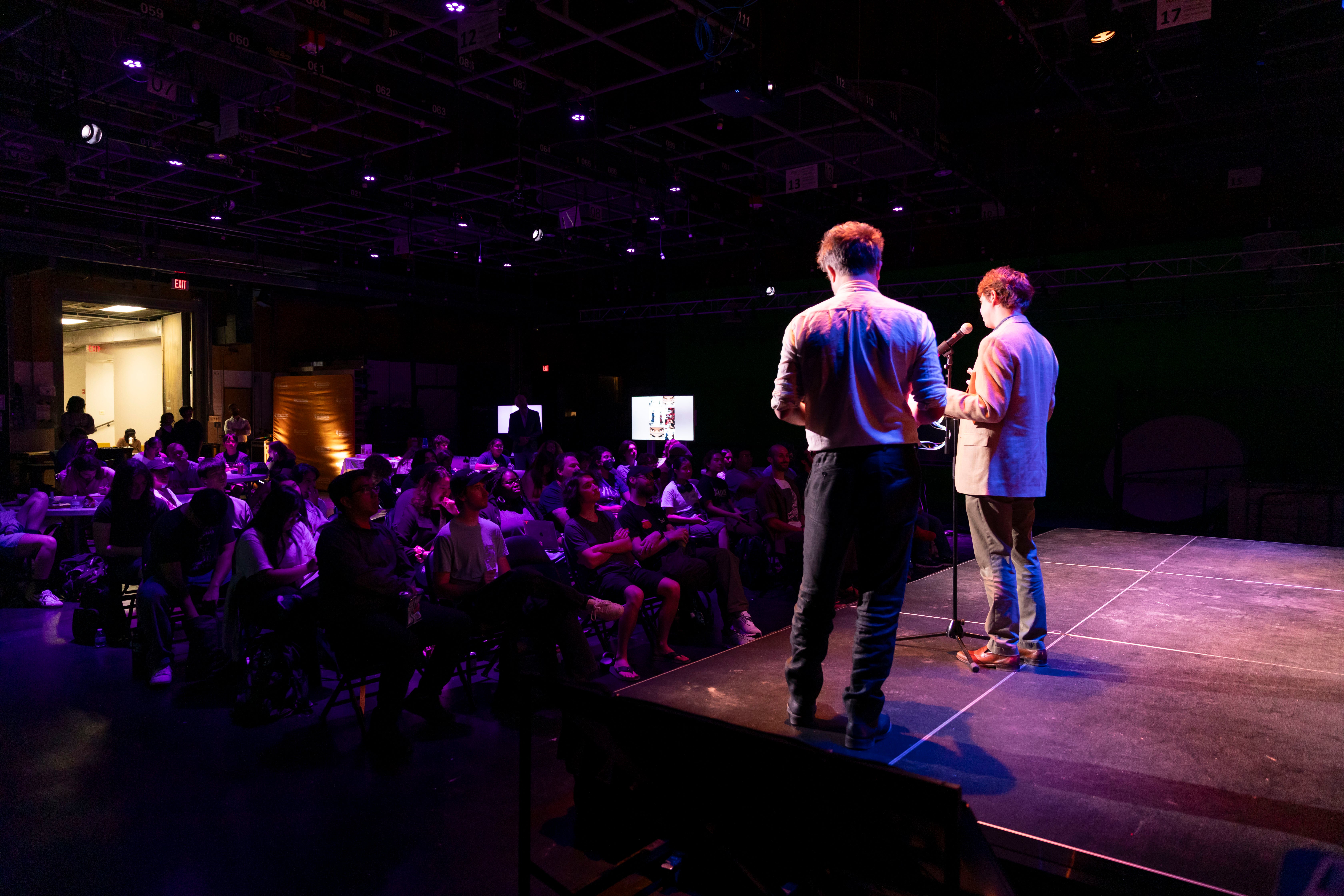 Two speakers stand on a stage and a crowd of students listens.