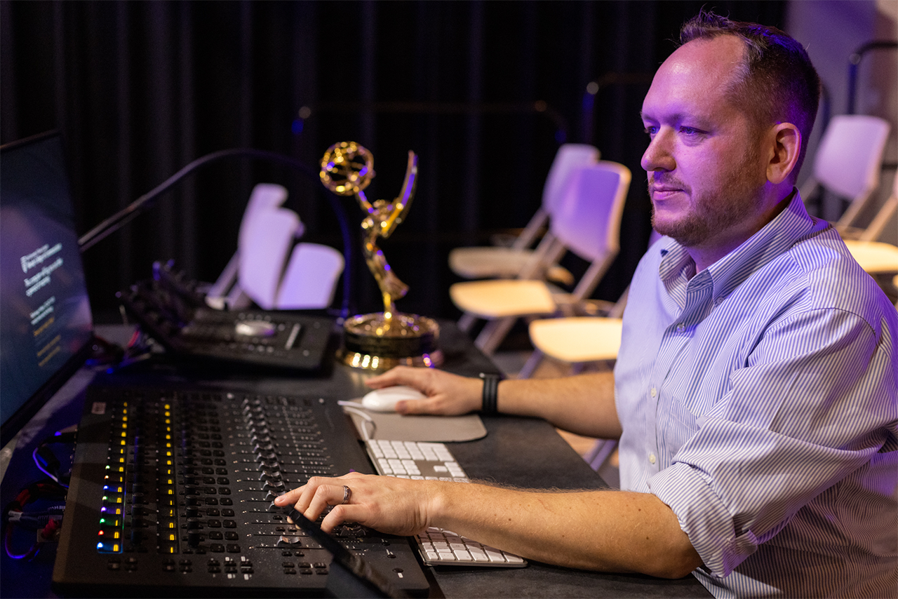 professor Korey Pereira sits at the computer editing audio with his emmy award on the desk