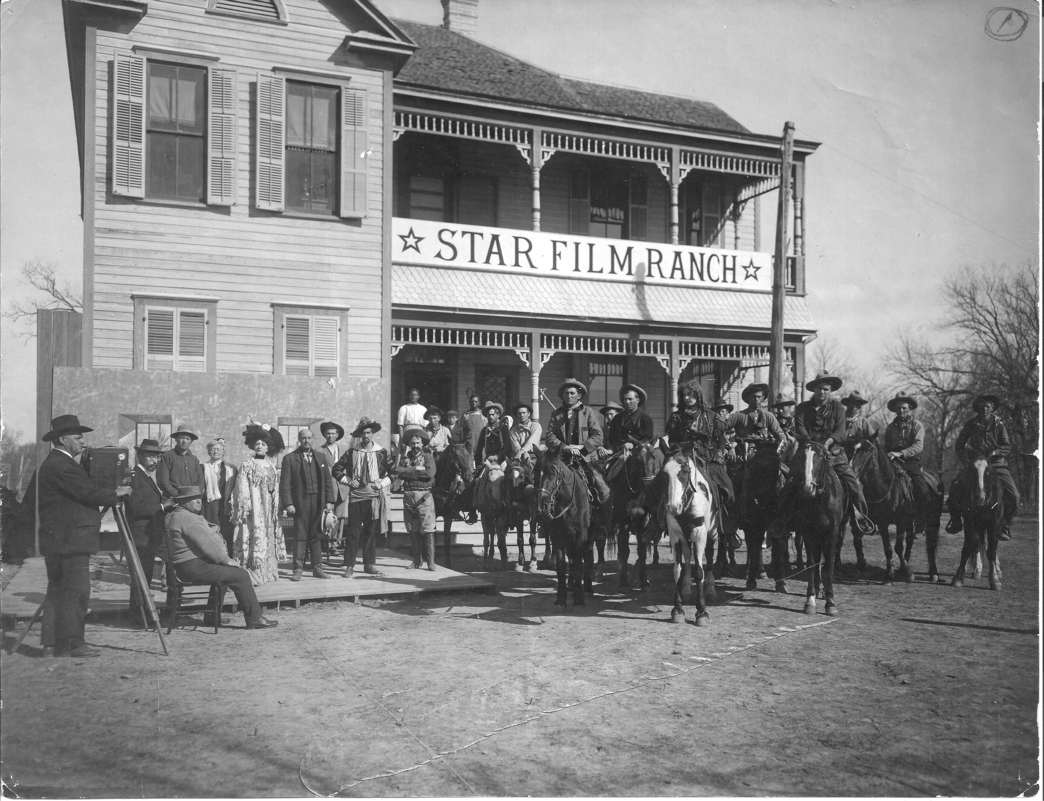 An old two-story house with the sign Star Film Ranch on the second floor balcony. Five horses and cowboys stand outside as well as other cast members for a film.