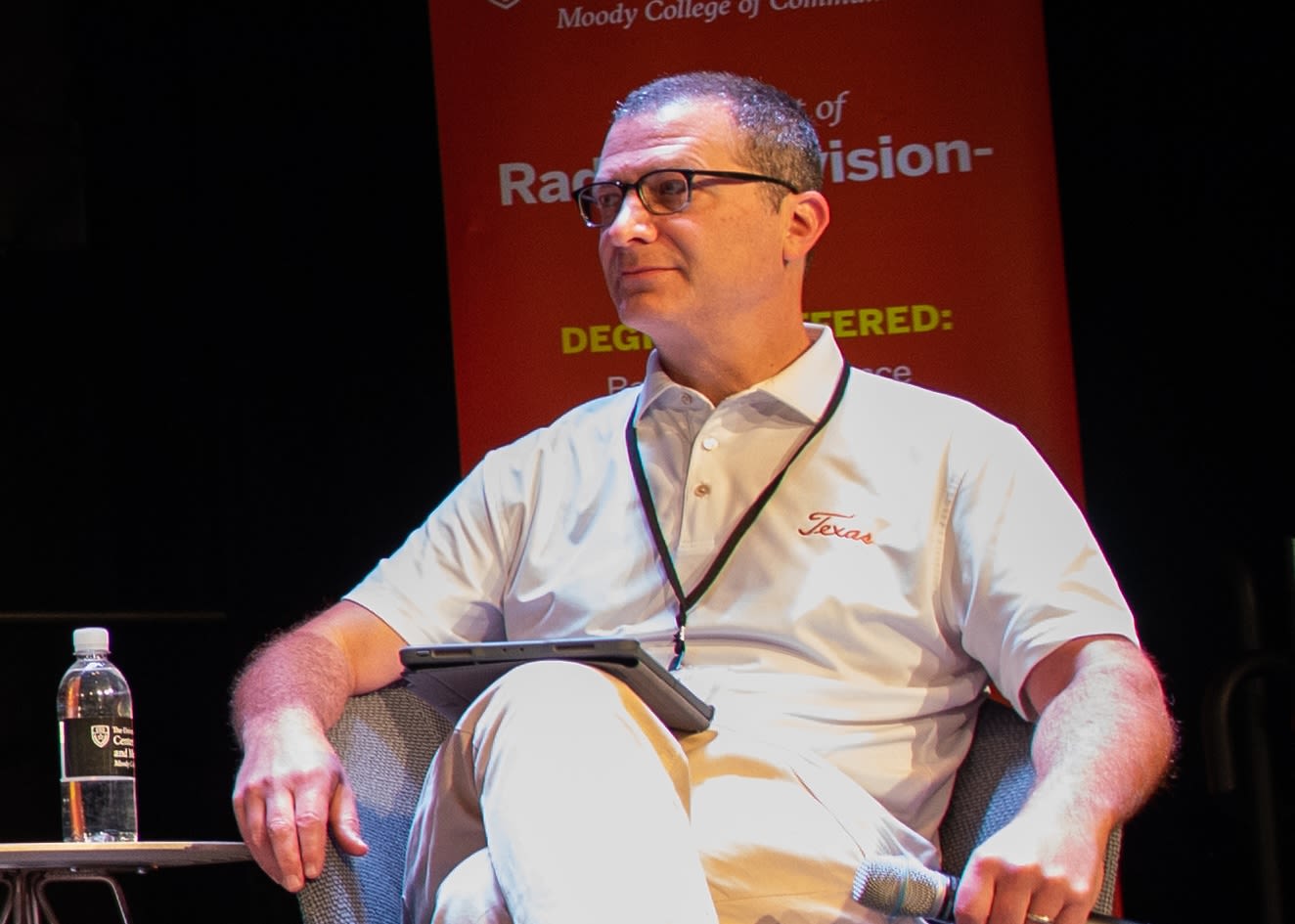 Man with glasses and lanyard sits with his legs crossed in a chair on stage.