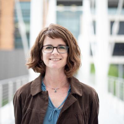 Photo of Dr. Maya Henry with a blurry background.  Short brown hair, rectangular glasses, blue shirt with a brown jacket.