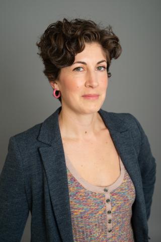 Headshot of Lara O'Toole, Sr. Program Coordinator at the CHC. White woman with short, curly brown hair wearing a dark gray blazer and multicolored tank top in front of a gray background