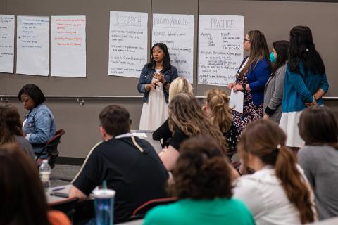 Attendees of the 2022 Health Communication Leadership Institute standing in front of large pieces of paper on the wall with writing on them presenting their group idea regarding a case study.