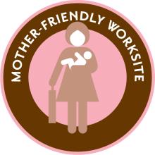 Pink circle with brown ring inside with overlay text reading "Mother-Friendly Worksite" and an illustration of a woman holding a briefcase and a baby.