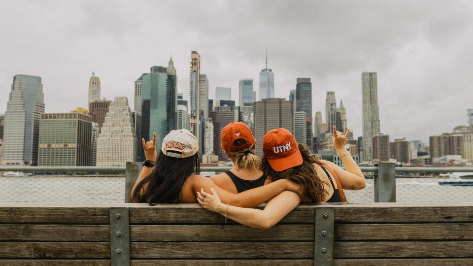 Three UTNY students wearing baseball caps sit on the a bench in Brooklyn overseeing the Manhattan skyline 