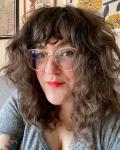 Kathy Cacace headshot, brown curly shoulder-length hair, wire glasses, gray v-neck t-shirt