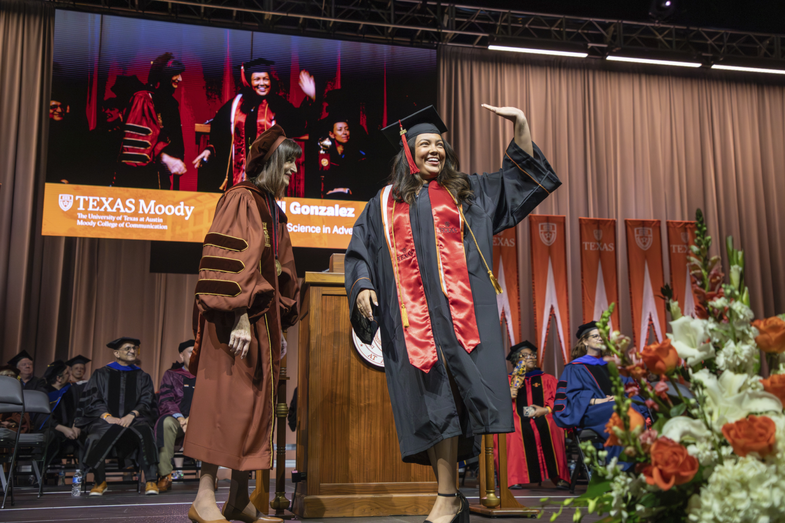 A smiling Moody College graduate waves to the audience while crossing the commencement stage.