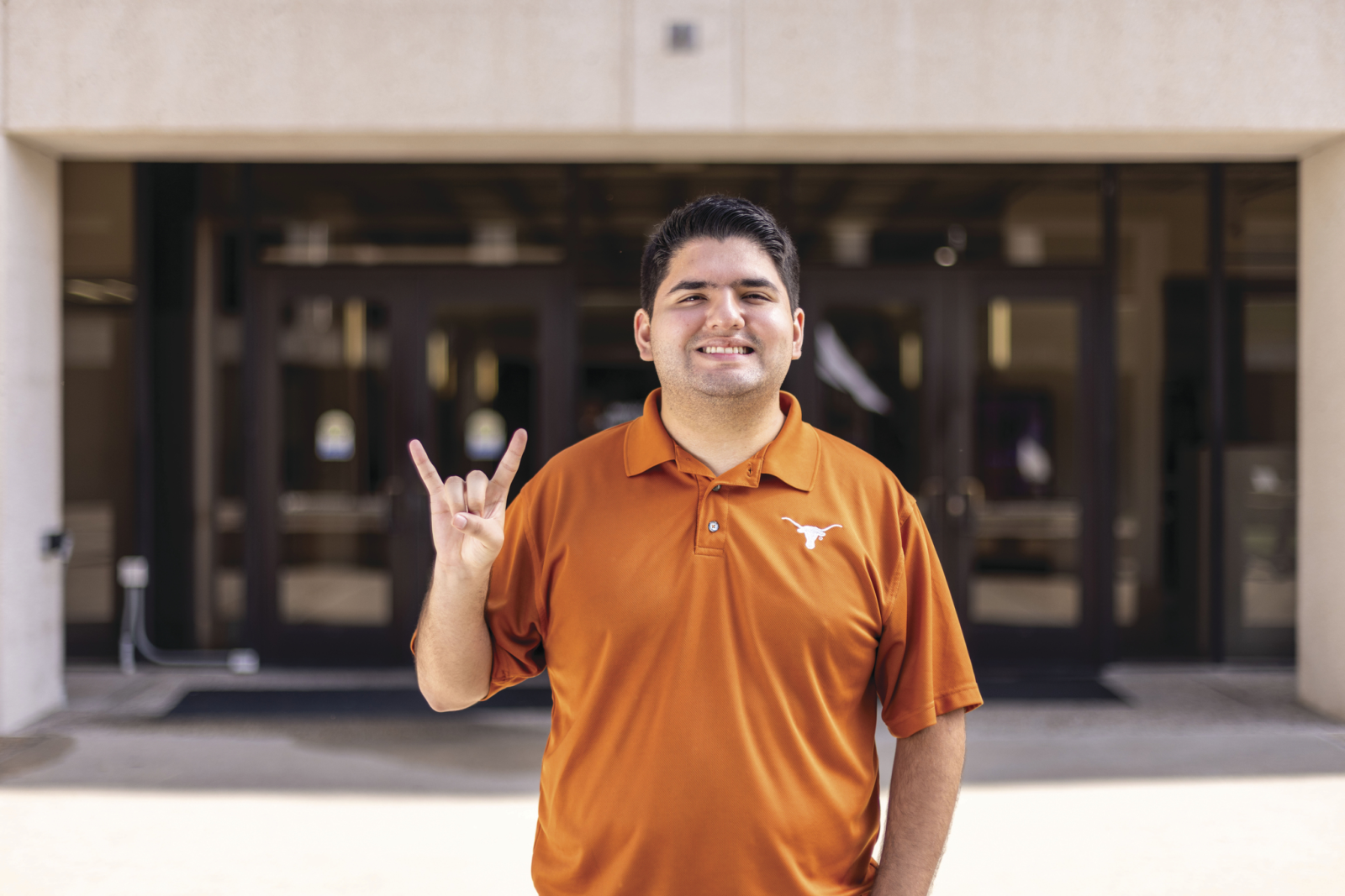 A student in an orange Longhorn polo smiles and makes the Hook ’em Horns gesture outside Moody College.