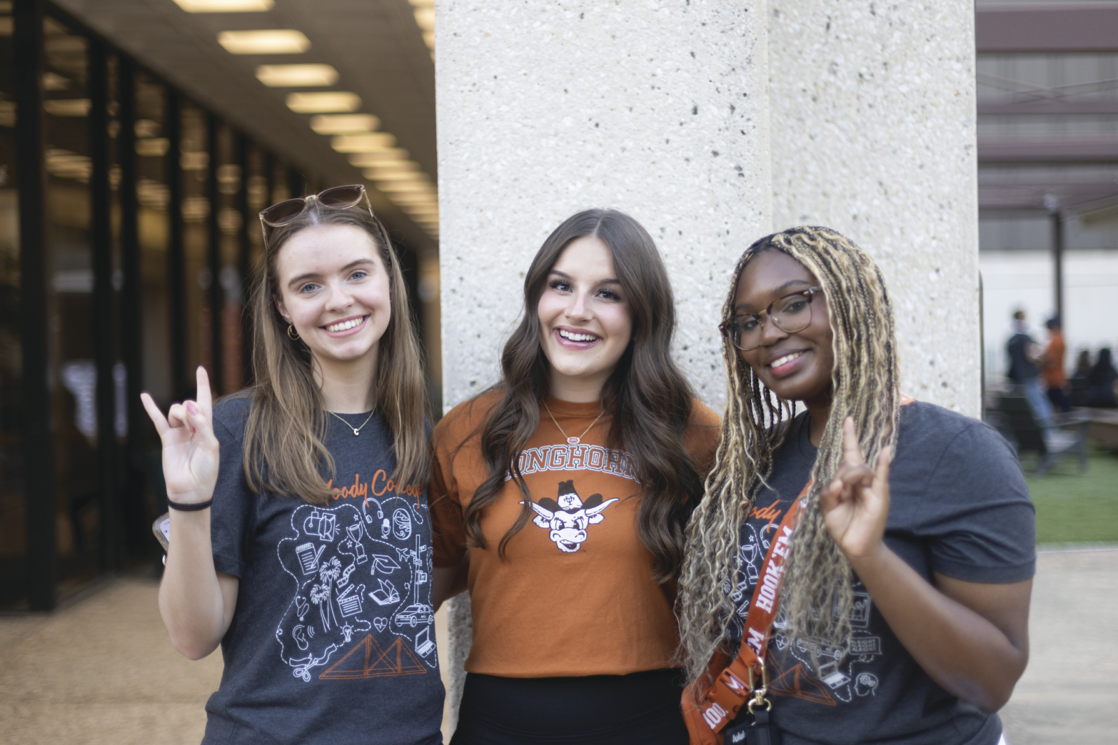 Three students smile together outside Moody College, with two making the Hook ’em Horns gesture.