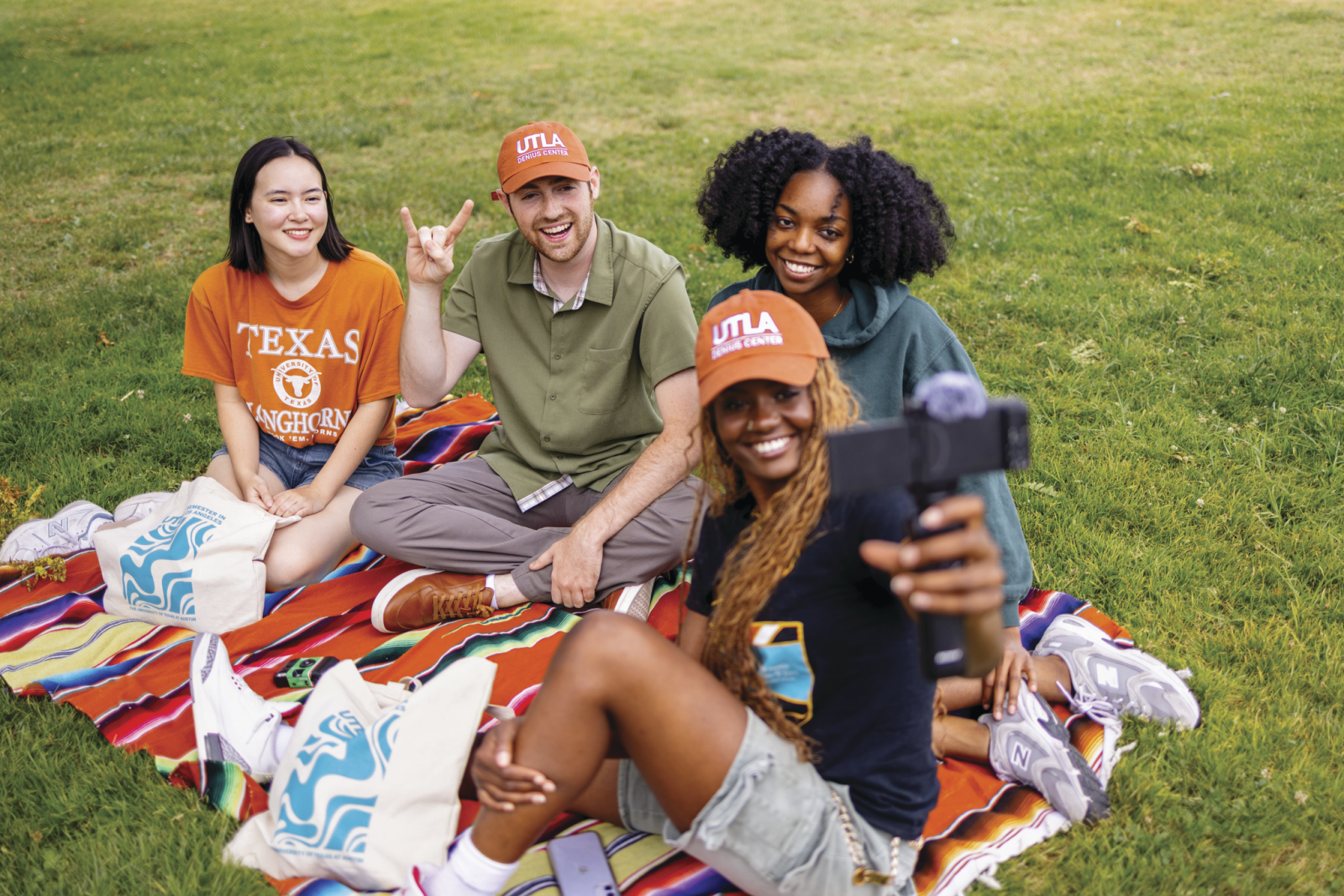 UTLA students sitting together on a picnic blanket, with one student making the Hook ’em Horns gesture and another filming the group.