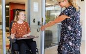 A woman holds a door open for another woman who is using a wheelchair