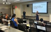 A presenter stands in front of a presentation and speaks to students and participants. 