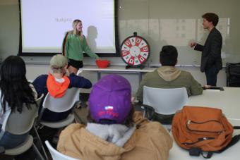 Students in a classroom facing front while two students play a game with a wheel 