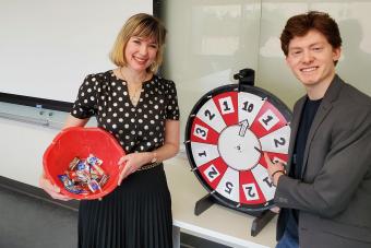A woman on the left holds a red bowl of candy and smiles toward the camera, next to her a young man holds a red and while wheel with numbers on it also smiling for the camera