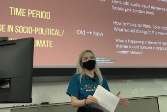 Student with a mask on holding papers talking in front of a screen with a slide on it
