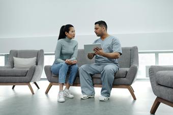 A woman and a man sitting on a gray couch facing each other. The man is showing the woman something on a tablet.