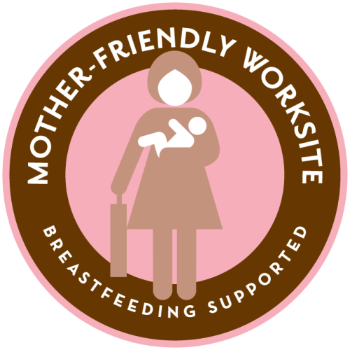 Pink circle with brown ring inside with overlay text reading "Mother-Friendly Worksite" and an illustration of a woman holding a briefcase and a baby.