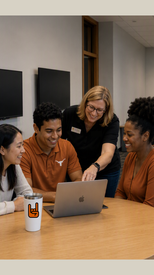 A group of people gathered around a laptop at a round table in a modern university-style collaboration space, while additional small groups work and converse at tables in the background. The room features neutral walls, overhead lighting, laptops, and an informal showcase or workshop environment focused on technology and teamwork.