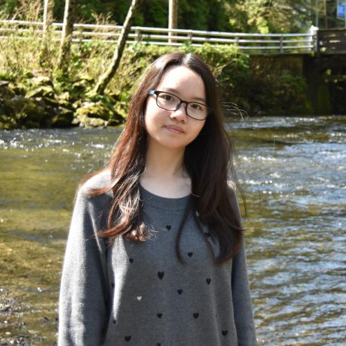 A young woman with dark hair and glasses stands in front of a river.