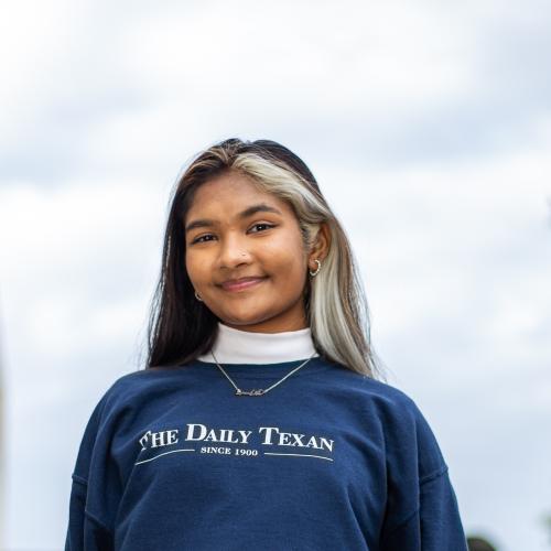 A young woman wears a blue Daily Texan sweatshirt. She has dark hair with one highlighted section.