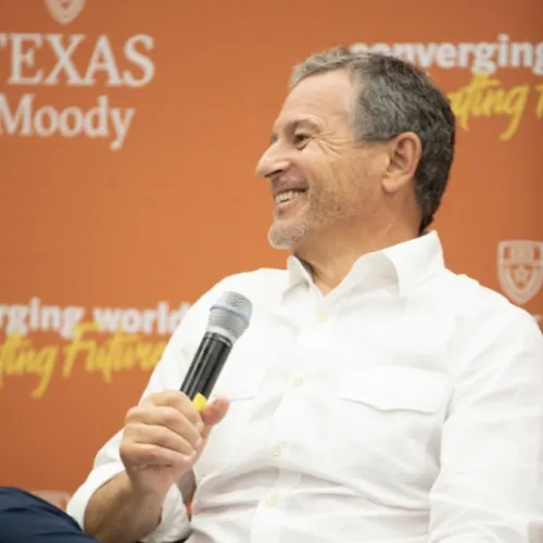 Bob Iger talking into a microphone, white shirt, orange background