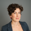 Headshot of Lara O'Toole, Sr. Program Coordinator at the CHC. White woman with short, curly brown hair wearing a dark gray blazer and multicolored tank top in front of a gray background