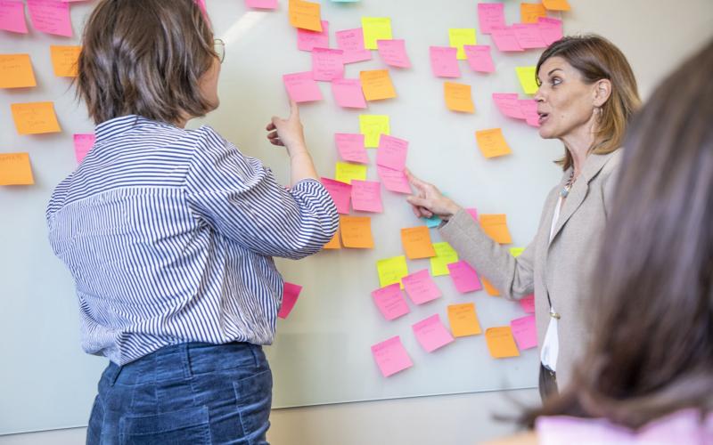 Two people talk in front of a whiteboard covered with sticky notes