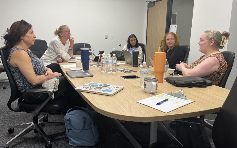 The image depicts a group of faculty in a conference room speaking and listening
