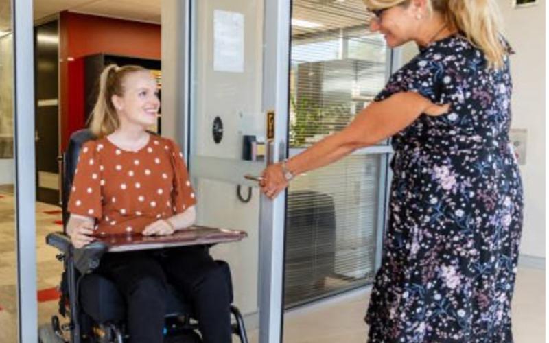 A woman holds a door open for another woman who is using a wheelchair